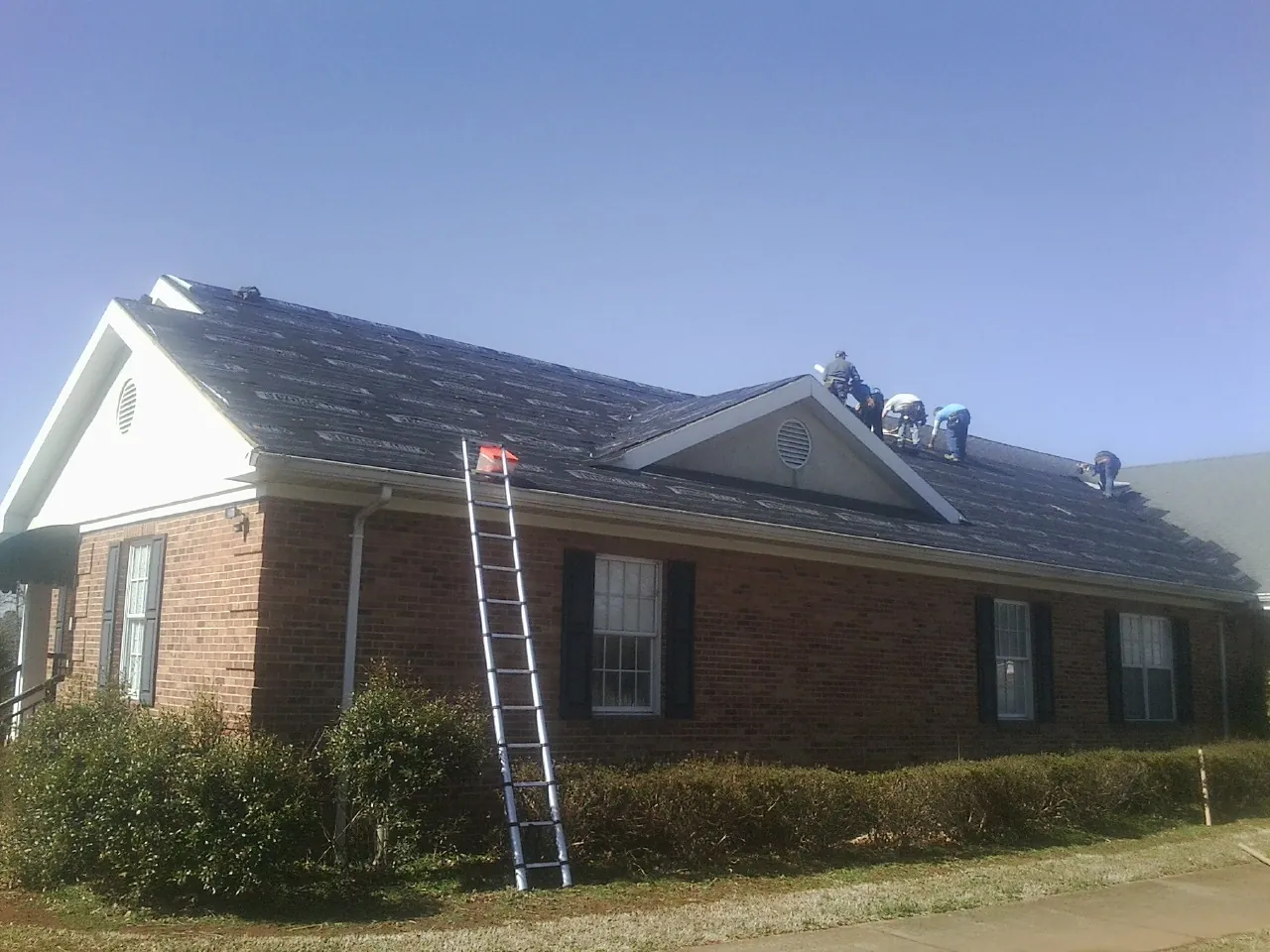 Crew tearing shingles off a brick ranch roof with a ladder set — Metal Roof Repair work by our crew in Childwold
