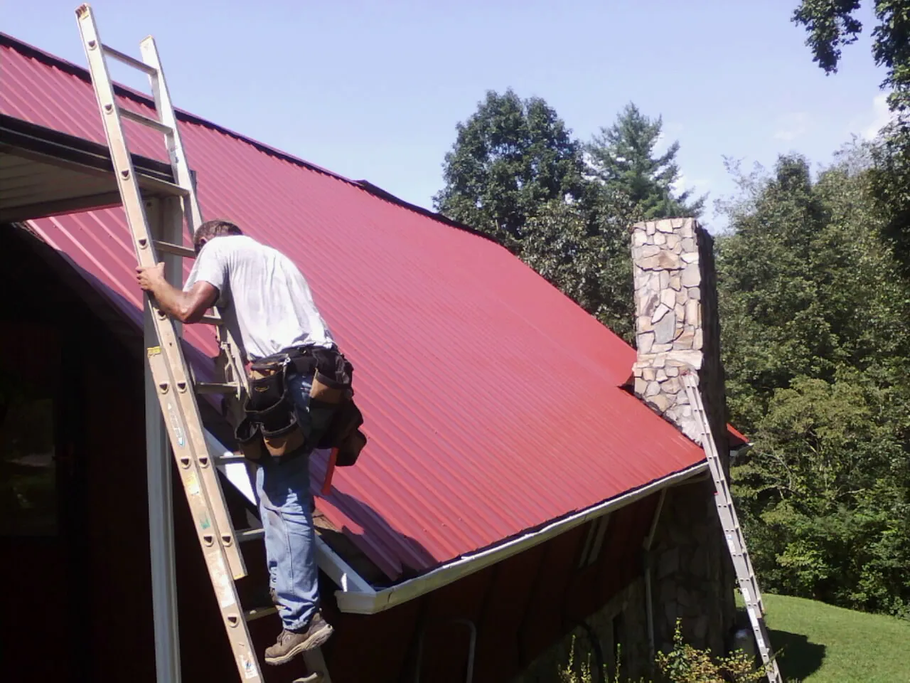 Roofer on a ladder beside a red metal roof and stone chimney — Soffit & Fascia Repair by Roofing Brothers in Childwold